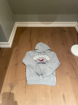 Grå hoodie från Hollister - Snygg grå hoodie från Hollister med tryck i vitt och vinrött framtill där det står 'Hollister California'. Klassisk huva och ribbade muddar. Perfekt för en avslappnad stil.