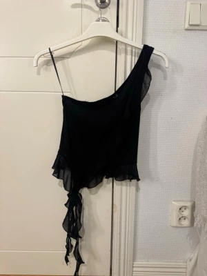 Svart oneshoulder topp med volang - Snygg svart oneshoulder topp med volangkant och asymmetrisk design. Tunt, lätt material med en axelbandslös sida och ett smalt axelband på andra sidan. Lång volangdetalj som hänger ner på ena sidan för extra edge.