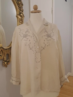 Krämvit blus med broderi - Snygg krämvit blus med ljusgrått blommigt broderi över bröstet och på ärmsluten. Blusen har knappar framtill, klassisk krage och trekvartsärmar. Materialet ser ut att vara mjukt och luftigt, perfekt för en avslappnad men stilren look.