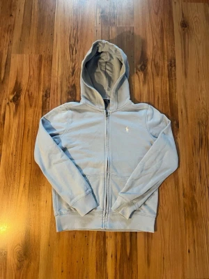 Ljusblå Ralph lauren zip hoodie  - Ljusblå Ralph lauren zip hoodie size 150 (12-14) Den är äkta och i mycket fint skick och sparsamt använd. Den mäter 54 cm i längd och 41 cm i bredd. Den har två knappt synbara minifläckar på vänstra ärmen, jag säger detta ifall någon undrar.                                                                  Hör av er om ni har några frågor kring tröjan!