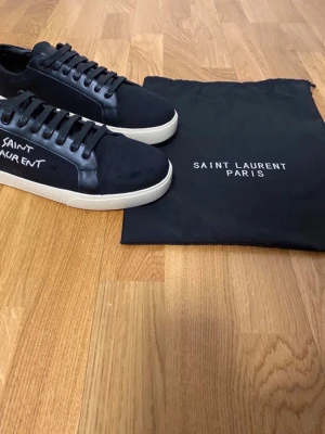Svarta Saint Laurent sneakers - Svarta sneakers från Saint Laurent med vit sula och handskriven logga på sidan. Skorna har snörning och detaljer i skinn samt canvasmaterial. Enkla men stilrena sneakers som passar perfekt till streetwear och modeintresserade.