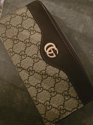 plånbok med GG-mönster - Snygg plånbok med klassiskt GG-mönster i beige och brunt. Plånboken har guldfärgad logga, dragkedja runtom och flera fack för kort och mynt. Perfekt accessoar för dig som gillar lyxiga detaljer och stilren design.