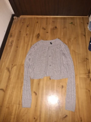 Grå cropped kofta från Divided - Grå stickad kofta från Divided med croppad passform och långa ärmar. Koftan har rund halsringning och knäppning framtill med små knappar. Perfekt att styla över en topp eller klänning.