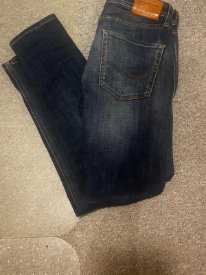 Mörkblå tapered jeans Jack & Jones - Snygga mörkblå jeans från Jack & Jones, modell Tapered/Erik. Klassisk femficksdesign med kontrastsömmar och en skön passform som smalnar av nedtill. Tillverkade i slitstarkt bomullsmaterial, perfekta för en avslappnad och trendig look.