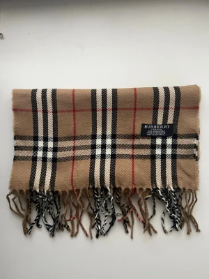 Burberry halsduk i kashmir - Klassisk halsduk från Burberry i 100% kashmir med ikoniskt rutmönster i beige, svart, vitt och rött. Halsduken har fransar i ändarna och är tillverkad i England. Perfekt accessoar för att lyfta din outfit.