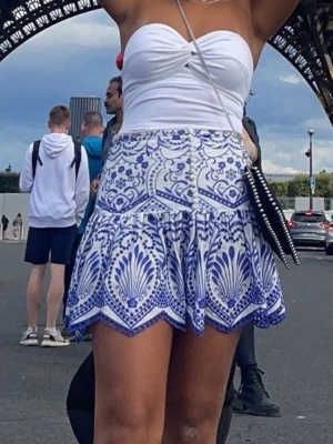 Vit och blå mönstrad kjol från Zara - Kort kjol från Zara i vitt och blått med ett unikt, detaljerat mönster. Kjolen har volangkant och hög midja, vilket ger en snygg och trendig siluett. Man kan bära kjolen lågmidjat även om man vill de. Perfekt för dig som vill sticka ut med en cool och somrig look. Kjolen är snålt använd. 