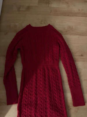 Röd stickad långärmad klänning - Mysig röd stickad klänning med kabelstickat mönster och långa ärmar. Klänningen har en rund halsringning och markerad midja, perfekt för kyligare dagar. Passformen är normal och materialet känns mjukt och varmt.