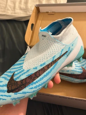 Nike blå fotbollsskor med mönster - Snygga fotbollsskor från Nike i ljusblått och vitt med stickad ovandel och stort brunt mönstrat swoosh på sidan. Skorna har snörfri design och ribbad struktur för bättre bollkontroll. Perfekta för dig som vill sticka ut på planen. Knappast andvända fick dom i gåva men de var lite för stora på mig men jag testade dom ändå och dom var jätte sköna men efteråt så rengjorde jag dom och glömde dom i mitt förråd. Säljer dessa för billigare än org pris som är ungefär 3200kr 