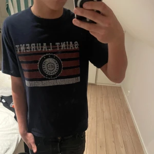 Mörkblå t-shirt från Saint Laurent - Snygg mörkblå t-shirt från Saint Laurent med tryck i rött och vitt. T-shirten är u extremt bra skick😃M men passar även S. Pris kan diskuteras vid snabb affär