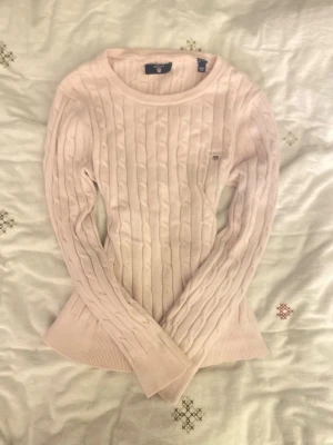 Ljusrosa kabelstickad tröja Gant - Supersnygg ljusrosa kabelstickad tröja från Gant med rund halsringning och diskret logga broderad på bröstet. Tröjan har långa ärmar och ribbade muddar vid ärmslut och nederkant. Perfekt för dig som gillar klassisk och stilren look.