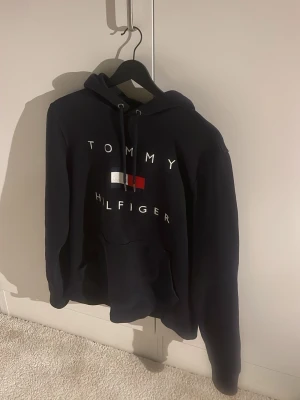 Mörkblå hoodie från Tommy Hilfiger - Snygg mörkblå hoodie från Tommy Hilfiger med klassisk logga och broderad text på bröstet. Hoodien har huva med dragsko och en stor magficka. Materialet är mjukt och skön. 