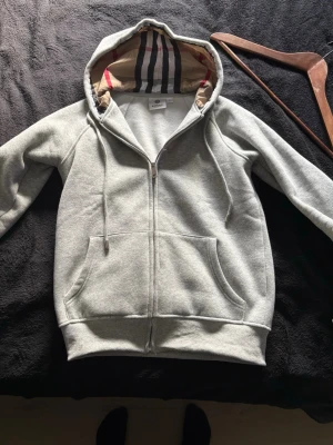 Grå zip-hoodie från Burberry - Stilren grå hoodie med dragkedja från Burberry. Klassisk huva med det ikoniska rutmönstret på insidan och broderad logga på bröstet. Mjuk bomullsblandning och känguruficka framtill. Perfekt för en avslappnad och trendig look.