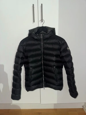 Colmar dam jacka - Snygg svart dunjacka från Colmar med huva och dragkedja framtill. Jackan har två fickor med dragkedja och Colmar-logga på ärmen. Perfekt för kalla dagar och har en klassisk pufferstil med horisontella sömmar.