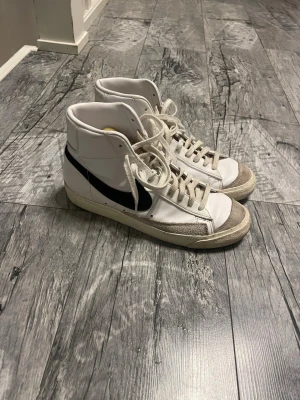 Nike Blazer Mid vita sneakers - Nike Blazer Mid sneakers i vitt skinn med grå mockadetaljer och svart swoosh på sidorna. Klassisk retrodesign med högt skaft och snörning. Perfekta för dig som gillar streetstyle och vill ha en ikonisk look.
