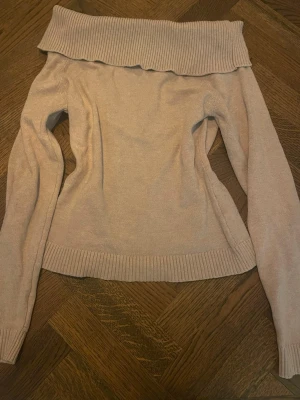 Beige off shoulder stickad tröja - Trendig beige stickad tröja med off shoulder-design och bred ribbad kant upptill. Långa ärmar och ribbade muddar vid ärmslut och nederkant. Perfekt för en snygg och avslappnad look.