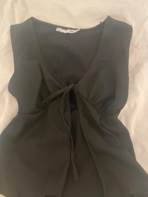Svart linne från lager storlek S - Snygg svart omlottopp med knyt framtill och v-ringad hals. Toppen är ärmlös och har en croppad passform som ger en trendig look. Perfekt att matcha med höga jeans eller kjol för en cool stil. Den är ifrån lager och är vi väldigt bra skick. Storlek S