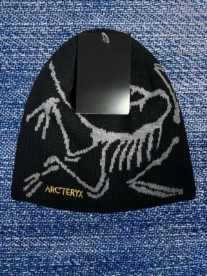 Arcteryx mössa - Håll värmen med stil i denna coola Arc'teryx mössa! Den är svart med ett iögonfallande grått skelettmönster som garanterat sticker ut. Guldfärgad Arc'teryx-logga nedtill på mössan ger en extra lyxig detalj. Perfekt för utomhusaktiviteter eller bara för att addera en unik touch till din vardagslook. En riktig statement-mössa för dig som gillar att synas!