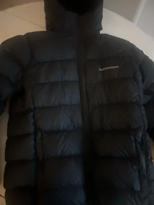 Svart dunjacka från Peak Performance - Säljer en svart dunjacka från Peak Performance med klassisk pufferdesign och huva. Jackan har dragkedja framtill och diskret logga på bröstet. Perfekt för kalla dagar och enkel att matcha med olika outfits.
