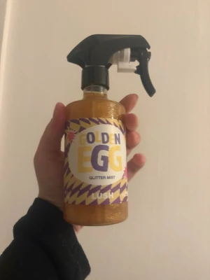 Golden Egg Glitter Mist från Lush - Golden Egg Glitter Mist från Lush är en ansiktsmist med guldig glittereffekt. Kommer i en genomskinlig plastflaska med spraymunstycke och har en färgstark etikett i lila och gult. Perfekt för att ge huden en skimrande finish.