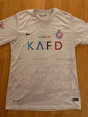 Vit Al Nassr Ronaldo matchtröja - Snygg vit Al Nassr matchtröja från Nike med Ronaldo och nummer 7 på ryggen. Tröjan har tryck i blått och rött framtill, klubbmärke på bröstet och flera sponsorloggor. Tillverkad i lätt och ventilerande Dri-FIT-material.