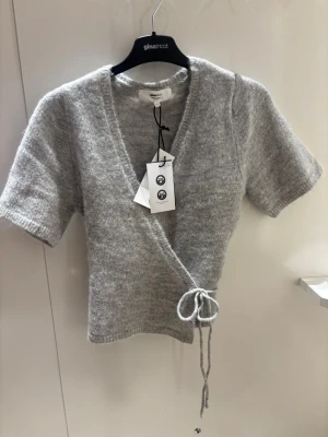 Grå omlottkofta från Gina Tricot - Mysig grå stickad omlottkofta från Gina Tricot i ullblandning. Kort modell med knytband i midjan och korta ärmar. Perfekt att styla över ett linne eller t-shirt för en trendig lager-på-lager-look.