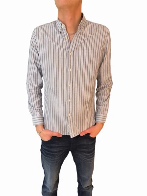 Randig skjorta med knappar - Snygg skjorta med smala vertikala ränder i vitt och grått. Klassisk krage, långärmad och med knäppning framtill. Perfekt för en stilren look och passar till jeans eller chinos.