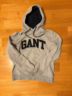 Grå hoodie från GANT - Snygg grå hoodie från GANT med stor svart logga broderad på bröstet. Mjuk insida och huva med mörkblått foder samt dragsnören. Perfekt för en avslappnad och trendig stil.