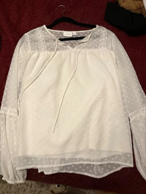 Vit prickig blus från Vila - Söt vit blus från Vila med transparenta långärmar och diskret prickigt mönster. Blusen har knytband vid halsen och spetsdetaljer på axlar och ärmslut. Tillverkad i ett lätt och luftigt material som ger en romantisk känsla. 