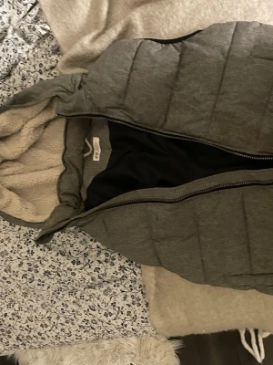 Grå dunväst med huva från KappAhl - Snygg grå dunväst från KappAhl med mysig fuskpälsfodrad huva och dragkedja framtill. Västen har en quiltad design och är perfekt för lager-på-lager-stil under kyliga dagar.