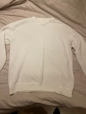 Vit stickad tröja med rund hals - Klassisk vit stickad tröja med rund halsringning och långa ärmar. Tröjan har ribbstickade muddar vid ärmslut och nederkant samt en diskret etikett nertill. Perfekt för lager på lager och enkel att matcha med olika stilar.