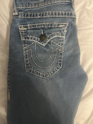 Blå True Religion jeans - Säljer ett par blå jeans från True Religion med deras ikoniska kontrastsömmar och stora bakfickor med lock och knapp. Klassisk rak passform och coola detaljer som sticker ut. Perfekta för dig som vill ha ett par jeans med attityd. En av knapparna där bak saknas.