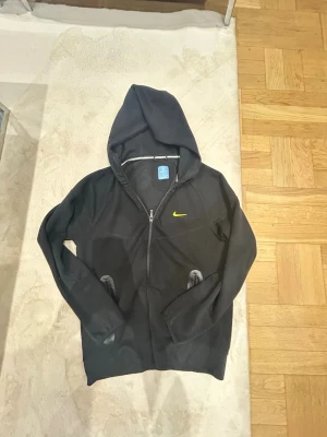 Svart Nike nocota kofta mycket bra Storlek S - Säljer en svart nike nocota från Nike med dragkedja och huva. Tröjan har två fickor framtill och en gul Nike-logga på bröstet.  Har använd Max 5-4 gånger Tillverkad i mjukt material som passar perfekt för en sportig och avslappnad stil. 100% bomull shippar samma dag samt köpet händer.