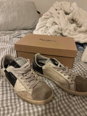 Pepe Jeans sneakers i beige och svart - Snygga sneakers från Pepe Jeans i beige och svart med vita detaljer. Skorna har snörning, rund tå och platt sula. Materialet är en mix av skinn och syntet. På hälen finns nitar som ger extra edge till looken. Perfekta för dig som gillar streetstyle.