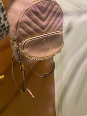 Rosa quiltad miniryggväska med kedjedetalj från Victoria secret - Söt rosa miniryggväska med quiltad framsida och guldfärgade kedjedetaljer. Väskan har en liten framficka med dragkedja och justerbara axelband. Perfekt för dig som vill ha en trendig och praktisk accessoar till vardagen. Oanvända 