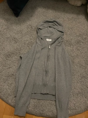  Gråa Hoodie och dragkedja - En grå hoodie från kidsbrandstorge med huva och hel dragkedja framtill. Västen är i mjukt, stickat material och har ribbade kanter nedtill. Perfekt att slänga över en t-shirt för en avslappnad look.