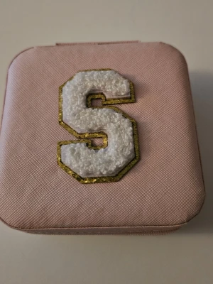 Rosa smyckesask med S-patch - Snygg rosa smyckesask med mjuk insida och fack för ringar och smycken. Locket pryds av ett vitt S i teddytyg med guldkant. Perfekt för att hålla ordning på dina accessoarer när du är på språng. Ca 10x10 cm