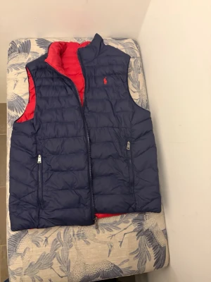 Vändbar dunväst från Polo Ralph Lauren - Snygg vändbar dunväst från Polo Ralph Lauren i marinblått och rött. Västen har två sidofickor med dragkedja och klassisk broderad logga på bröstet. Perfekt för lager på lager och enkel att matcha med olika outfits.