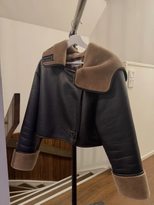 Svart croppad skinnjacka med teddykrage - Snygg croppad svart skinnjacka med bred beige teddykrage och matchande muddar. Jackan har oversized passform och stora slag, vilket ger en cool look. Perfekt för dig som vill sticka ut med en trendig jacka i höst och vinter.