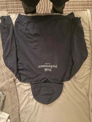 Mörkblå hoodie från Peak Performance - Mörkblå hoodie från Peak Performance med broderad logga i vitt på bröstet. Klassisk känguruficka framtill och huva med dragsko. Perfekt för en avslappnad och sportig stil. Den har en vit fläck bredvid logan.