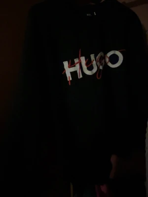 Svart sweatshirt från Hugo - Cool svart sweatshirt från Hugo med stor vit och röd logga framtill. Tröjan har rund halsringning och långa ärmar. Perfekt för dig som gillar streetwear och vill ha en enkel men snygg look.