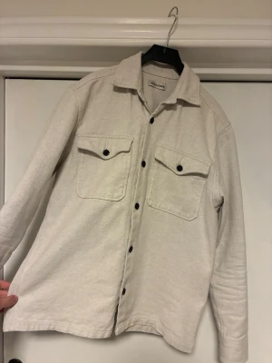 Beige overshirt  - Stilren beige overshirt från Jack and Jo es med svarta knappar och två bröstfickor. Skjortjackan har klassisk krage och är långärmad, perfekt att bära över en t-shirt för en avslappnad look.