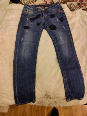 Blå jeans med patches och slitningar - Säljer ett par blå jeans med coola svarta patches och slitningar framtill. Jeansen har raka ben och klassisk femficksmodell. Materialet är denim och de har en avslappnad streetstil med unika detaljer som stjärnor och figurer på framsidan.