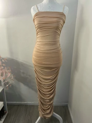 Klänning - Supersnygg beige långklänning med smala axelband och figurnära passform. Klänningen har rynkade detaljer längs hela kroppen och en dragkedja i ryggen. Storlek XS men passar även S i och med att den är så pass stretchig. Aldrig använd, endast testad. 🤎