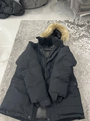 Svart dunjacka med päls från Everest - Svart dunjacka från Everest med huva som har en snygg pälsdetalj. Jackan har stora fickor med knappar, dragkedja framtill och är fodrad för extra värme. Perfekt för kalla dagar och har en klassisk, stilren look.