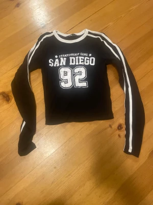 Svart långärmad tröja FB Sister XS - Cool svart långärmad tröja från FB Sister i storlek XS med vita ränder längs ärmarna och tryck på bröstet med texten 'San Diego 92'. Rund halsringning med vit kant. Perfekt för dig som gillar sportig och avslappnad stil.