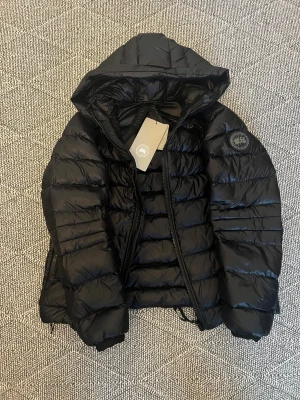 Svart dunjacka från Canada Goose - Säljer en svart dunjacka från Canada Goose med huva och snygga horisontella sömmar. Jackan har dragkedja framtill och logotyp på ärmen. Perfekt för kalla dagar och har en modern, puffad look som är riktigt trendig.