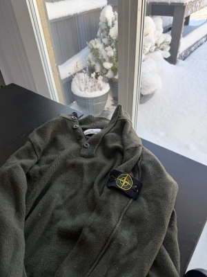 Grön stickad tröja från Stone Island - Snygg grön stickad tröja från Stone Island med klassisk patch på ärmen. Tröjan har ribbad krage, knappslå med fyra knappar och en dragkedja vid halsen. Perfekt för dig som gillar streetwear och vill ha något som sticker ut.