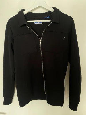 Svart overshirt från Jones med dragkedja - Svart overshirt från Jones med stilren design, två bröstfickor och hel dragkedja framtill. Jackan har klassisk krage och långärmade ärmar. Perfekt för dig som gillar minimalistisk och clean look.