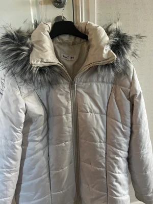 Vit pufferjacka med fuskpäls - Snygg vit pufferjacka med fluffig huva och grå fuskpälsdetaljer. Jackan har quiltad design, dragkedja framtill och långärmade ärmar. Perfekt för kalla dagar med sin varma och stilrena look.