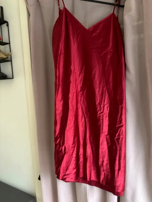 Röd satin klänning med öppen rygg - Snygg röd klänning i satin med smala axelband och v-ringad hals. Klänningen har en öppen rygg med knytband för extra detalj. Den är lång och har en slät, glansig yta som ger en lyxig känsla. Skulle räkna de som nattplagg å inte en klänning att ha ute. Ganska stor i storlek